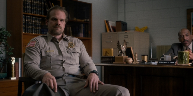 Wide shot frame from Stranger Things — sitting, desk, uniform