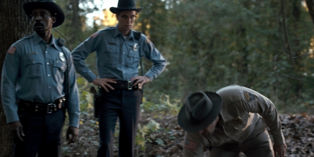 Wide shot frame from Stranger Things — standing, hat, gun