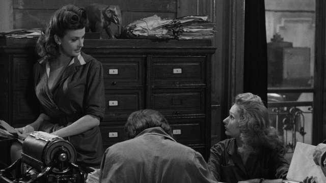 Wide shot frame from Léon Morin, prêtre — sitting, book, standing