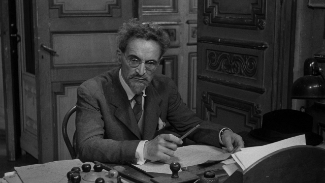 Wide shot frame from Léon Morin, prêtre — sitting, book, door