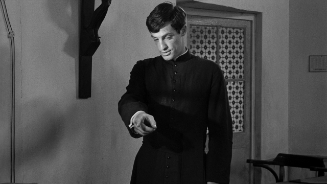 Wide shot frame from Léon Morin, prêtre — standing, room, hand