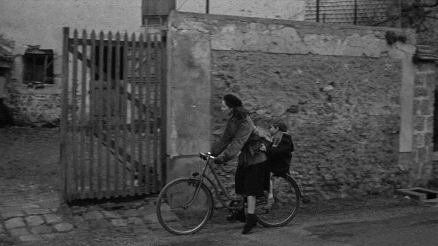 Wide shot frame from Léon Morin, prêtre — sitting, street, hat