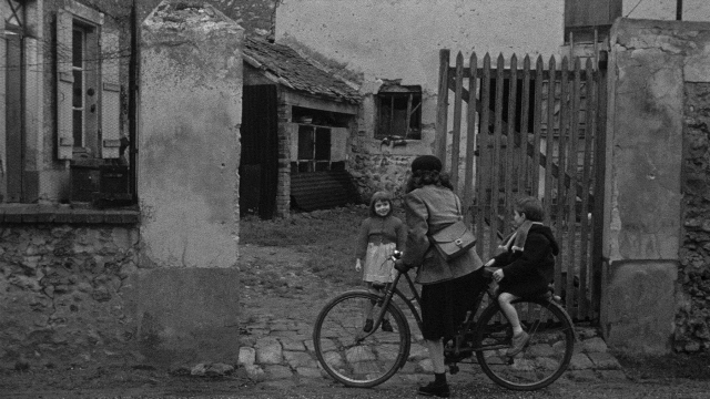 Wide shot frame from Léon Morin, prêtre — standing, street, hat