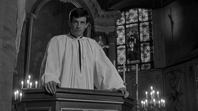 Wide shot frame from Léon Morin, prêtre — standing, tie, face
