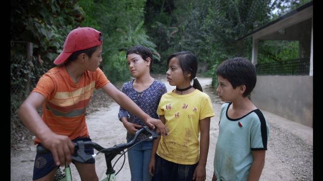 Wide shot frame from Noche de Fuego — standing, hat, bicycle