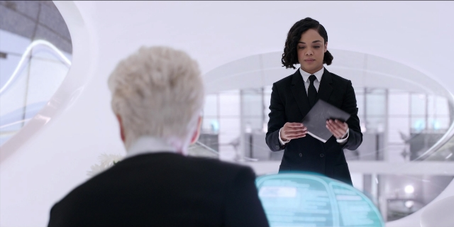 Wide shot frame from Men in Black International — sitting, standing, suit