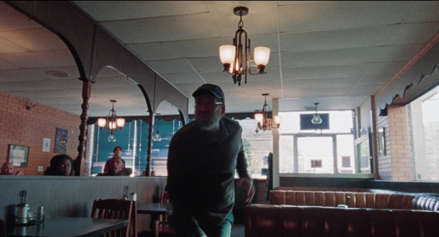 Wide shot frame from Dream Scenario — sitting, standing, table