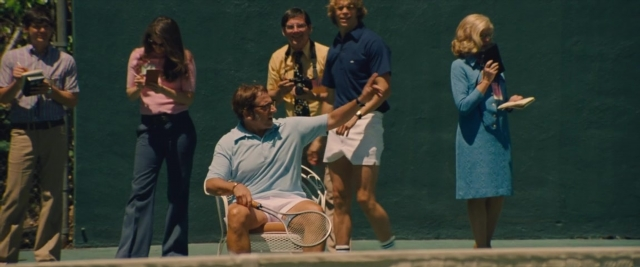 Wide shot frame from Battle of the Sexes — sitting, book, standing