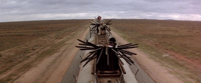 Wide shot frame from Mad Max 2 — sitting, street, car