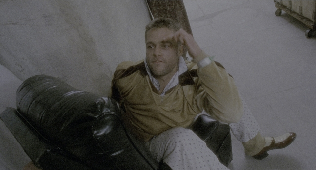 Wide shot frame from Twelve Monkeys — sitting, room, hand