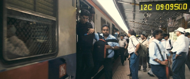 Wide shot frame from The Lunchbox — standing, phone, train