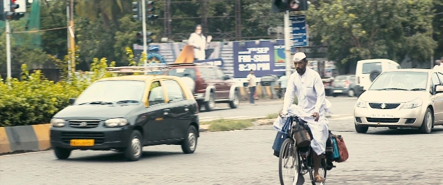 Wide shot frame from The Lunchbox — street, hat, car
