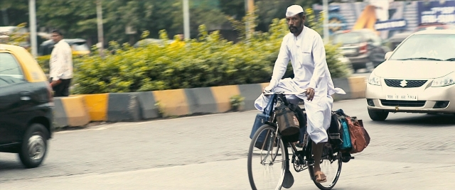 Wide shot frame from The Lunchbox — street, hat, car