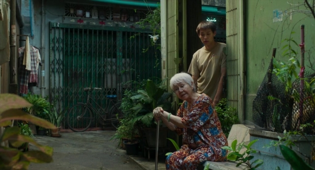 Wide shot frame from Lahn Mah — sitting, standing, hand