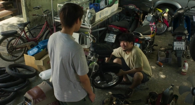 Medium shot frame from Lahn Mah — standing, table, bicycle