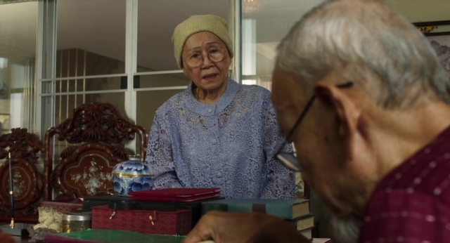 Wide shot frame from Lahn Mah — sitting, table, dress