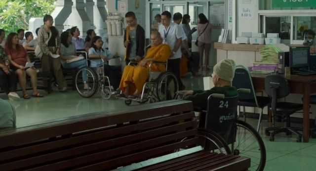 Wide shot frame from Lahn Mah — sitting, desk, standing