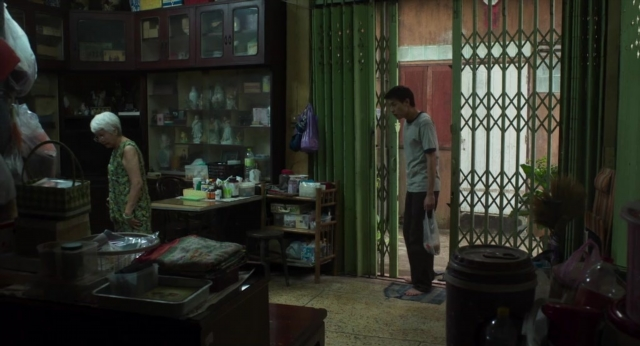 Medium shot frame from Lahn Mah — standing, table, hand