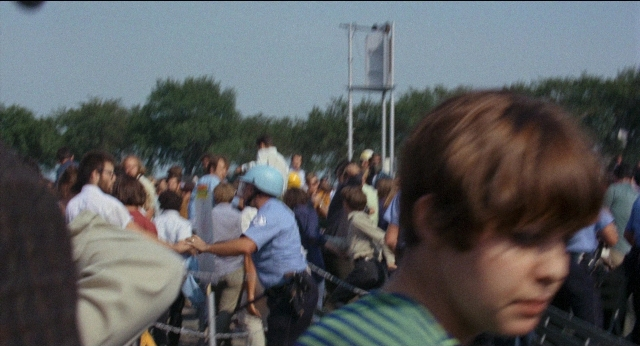 Frame from Medium Cool — standing, dialogue, hat