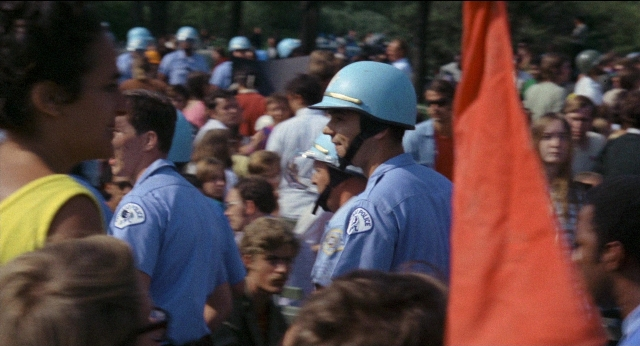 Close-up frame from Medium Cool — standing, dialogue, hat