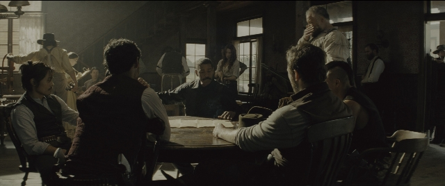 Wide shot frame from The Magnificent Seven — sitting, book, standing