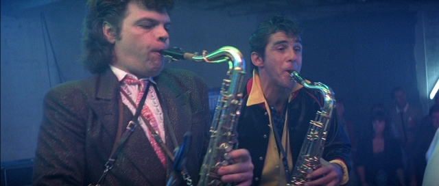 Wide shot frame from The Adventures of Buckaroo Banzai Across the 8th Dimension — room, hair, saxophone