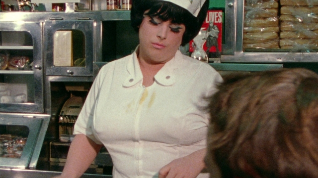 Wide shot frame from Female Trouble — standing, dialogue, hat