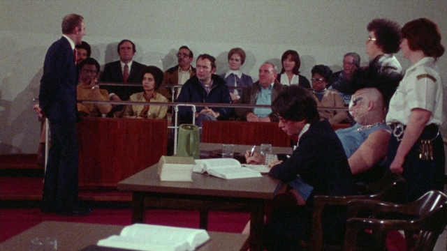 Wide shot frame from Female Trouble — sitting, book, standing