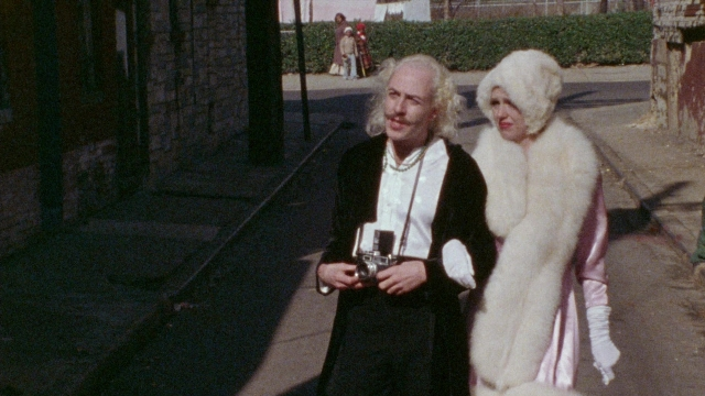 Wide shot frame from Female Trouble — walking, street, coat