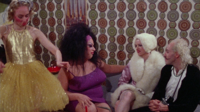 Wide shot frame from Female Trouble — sitting, room, coat