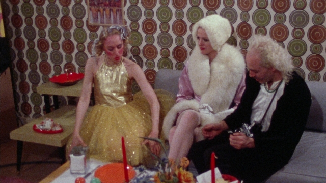 Medium shot frame from Female Trouble — sitting, plate, room