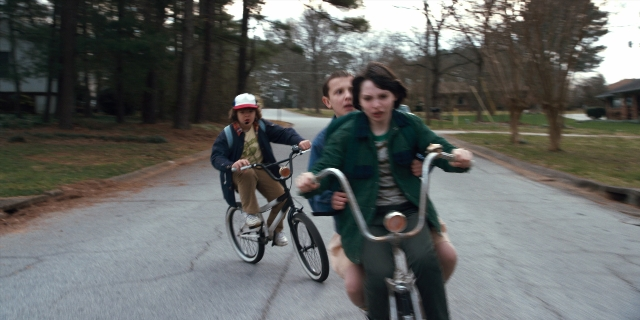 Wide shot frame from Stranger Things — street, hat, bicycle