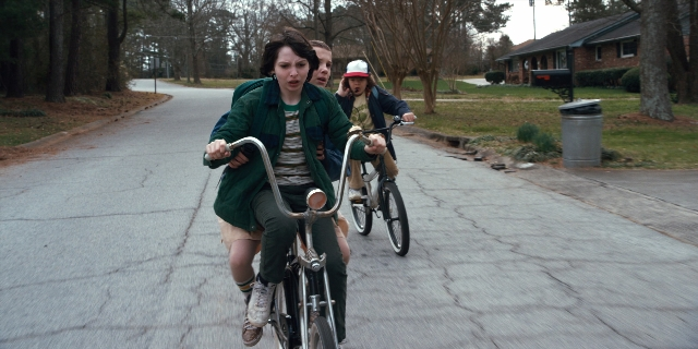 Wide shot frame from Stranger Things — street, hat, bicycle