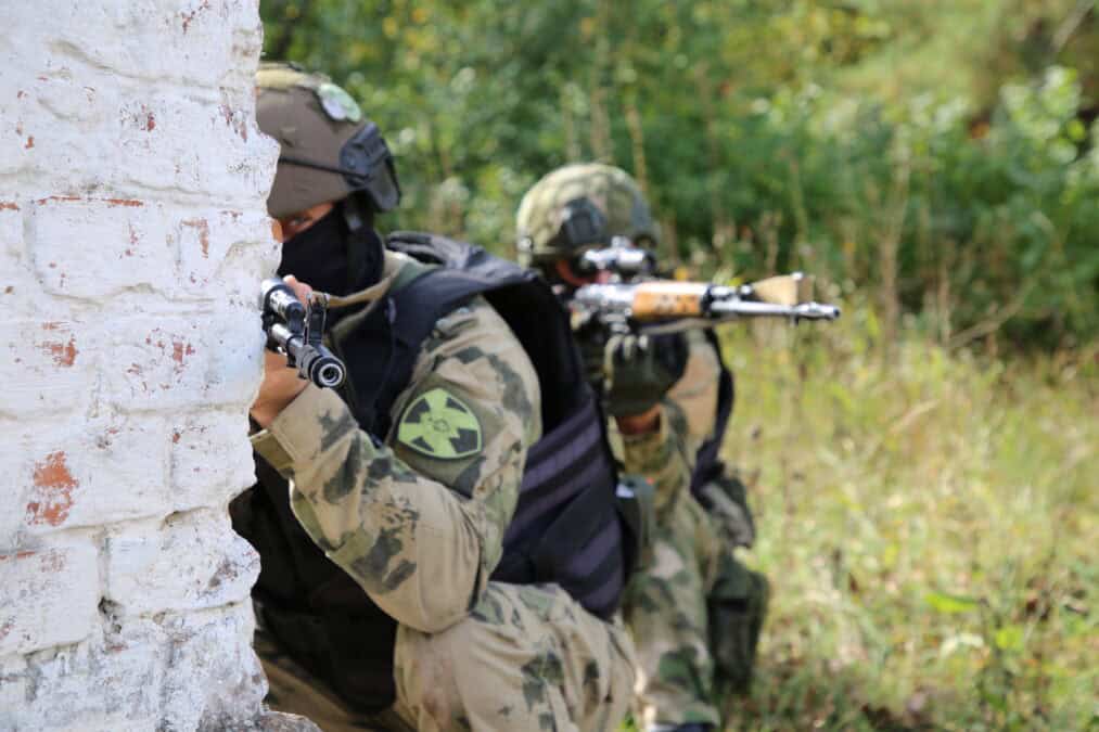 Двое вооружённых бойцов укрываются у кирпичной стены.