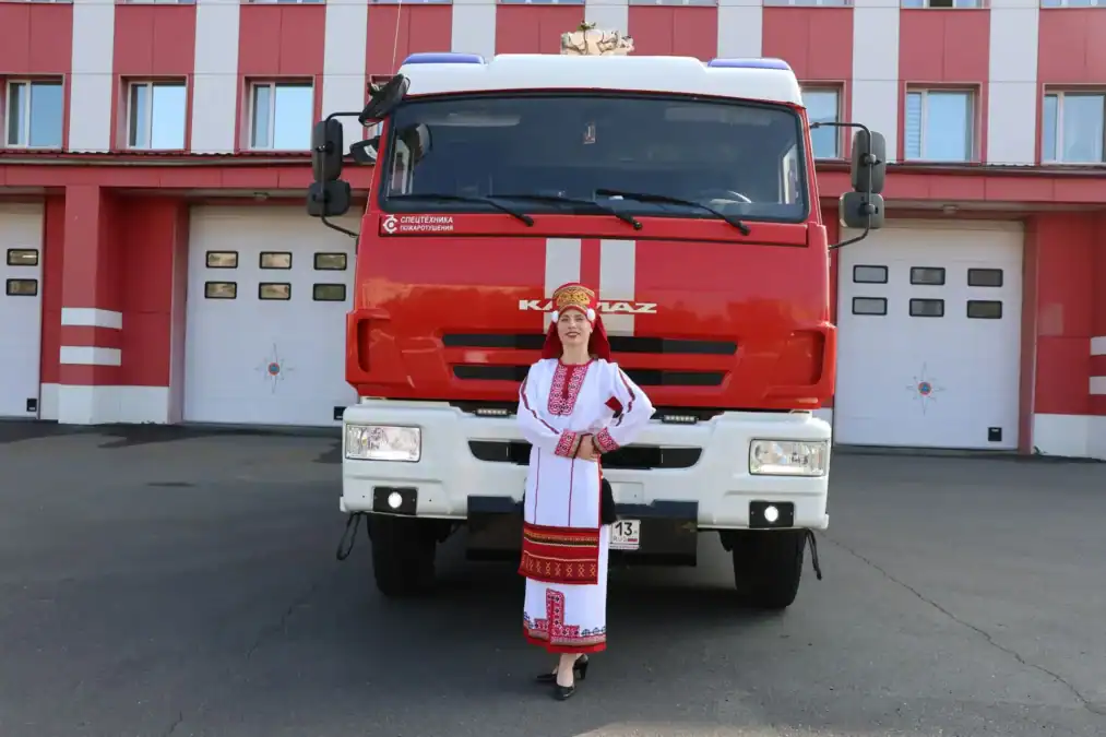 Женщина в национальном костюме стоит перед пожарной машиной у здания части.