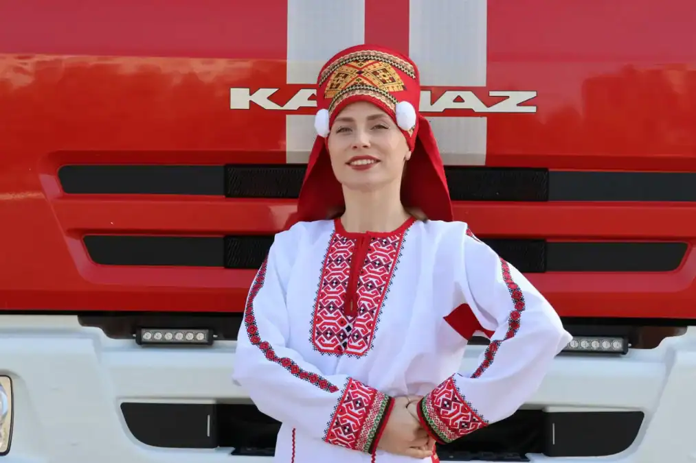 Женщина в национальном костюме стоит перед красным автомобилем КАМАЗ.