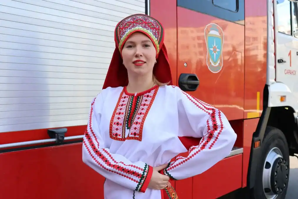 Женщина в вышитом национальном костюме стоит у пожарного автомобиля.
