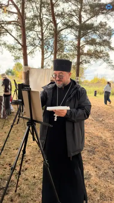 Священник рисует на мольберте на открытой площадке среди деревьев.