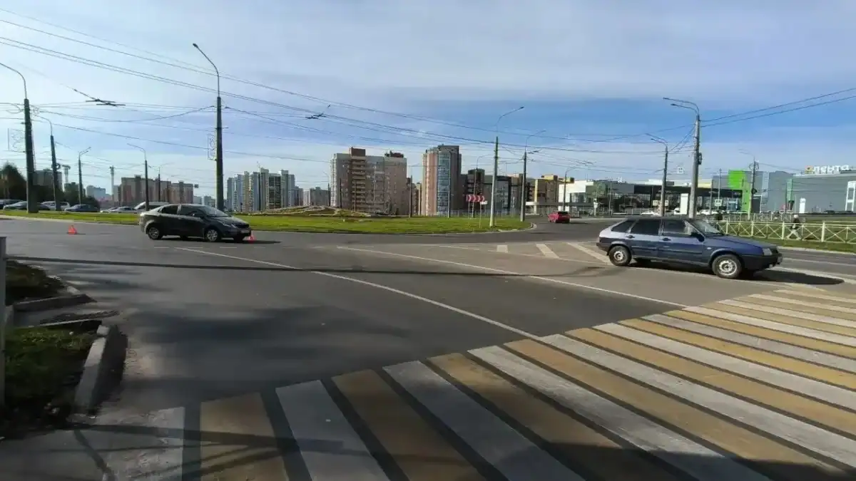 Автомобили проезжают через городской перекрёсток с пешеходным переходом и аварийным конусом.