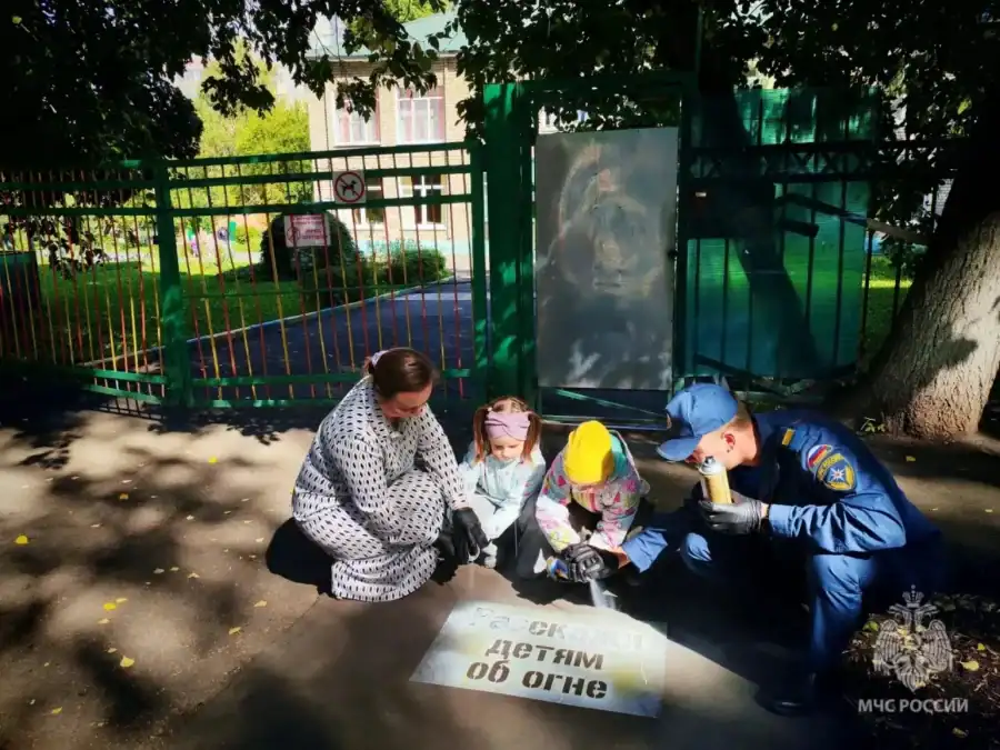 Женщина, дети и сотрудник МЧС сидят у трафарета с предупреждением не оставлять детей одних.