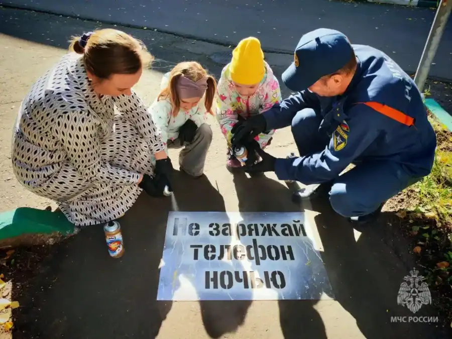 Женщина, двое детей и сотрудник МЧС наносят предупреждающую надпись на асфальт через трафарет.