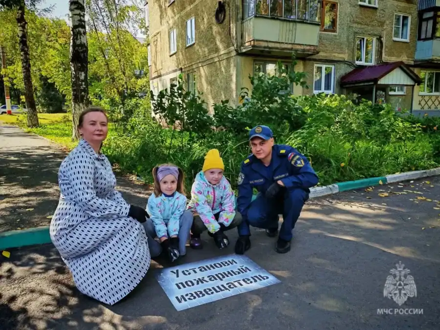 Женщина, двое детей и сотрудник МЧС сидят у надписи об установке пожарного извещателя.