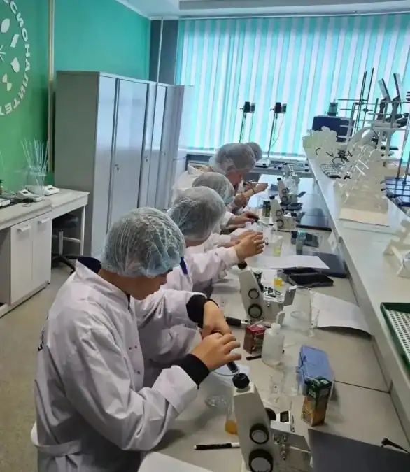 Учащиеся в белых халатах и защитных сеточках работают с микроскопами в лаборатории.