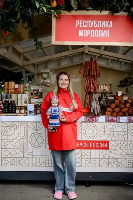 Женщина держит куклу у стенда с копчёностями и надписью «Республика Мордовия».