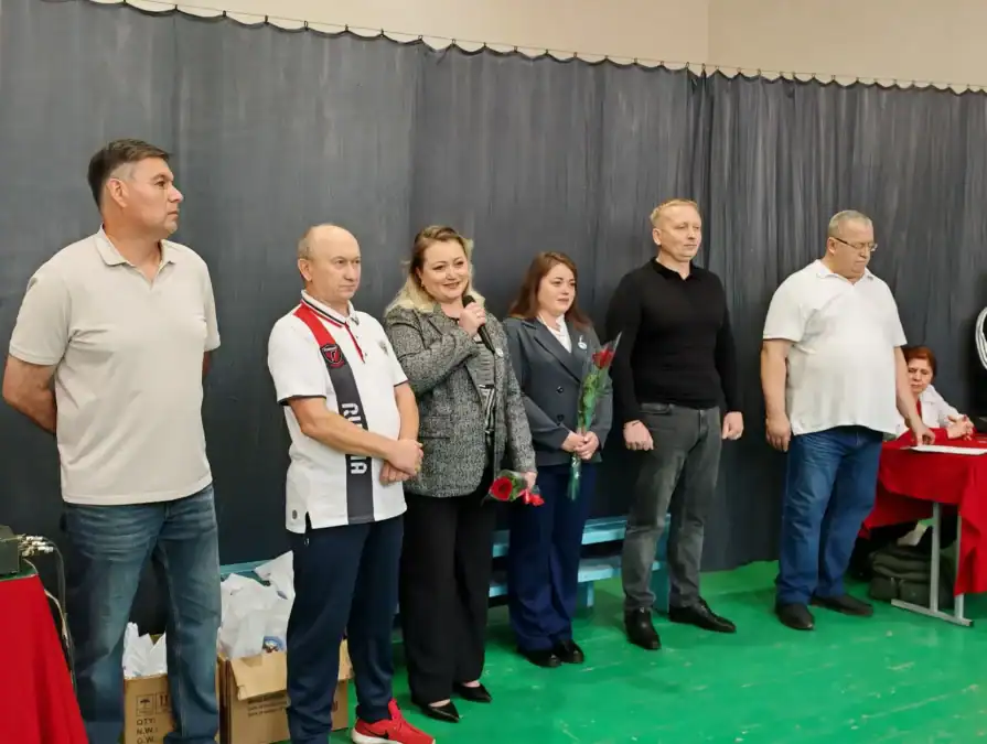 Шесть взрослых стоят у стены спортивного зала рядом со столом.
