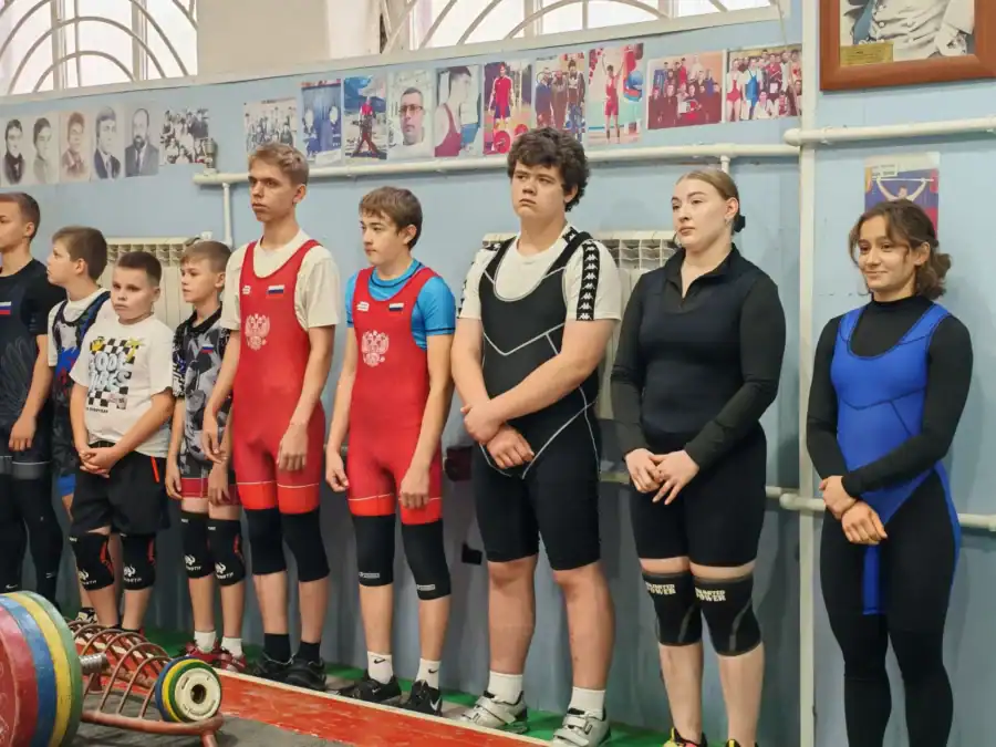 Юные спортсмены в тяжелоатлетической экипировке стоят вдоль стены зала.