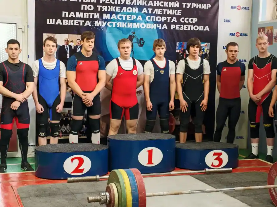 Тяжелоатлеты стоят за пьедесталом перед баннером соревнований.