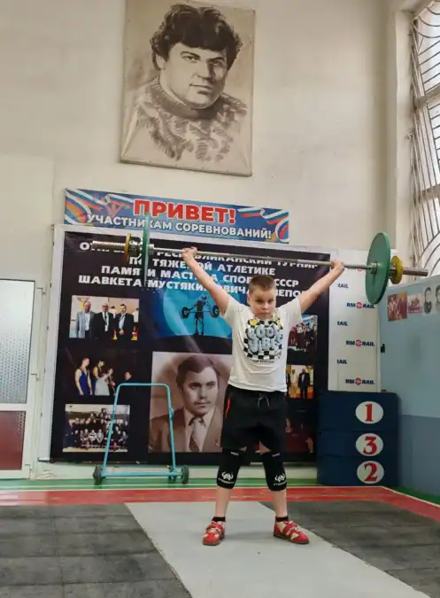 Юный спортсмен поднимает штангу над головой на помосте.