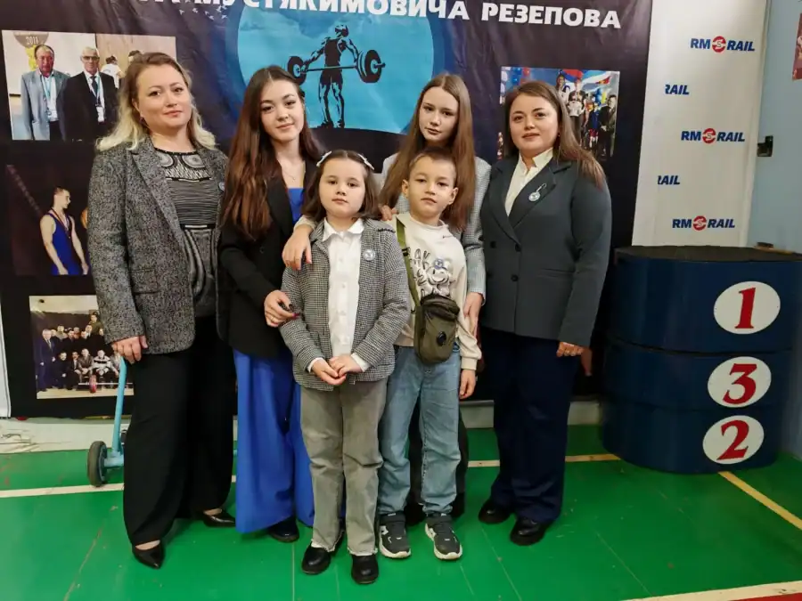 Женщины и двое детей позируют у пьедестала в спортивном зале.