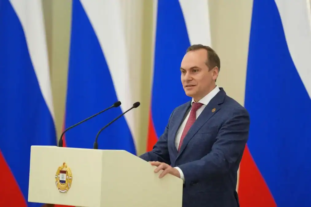 Мужчина выступает за трибуной на фоне российских флагов.
